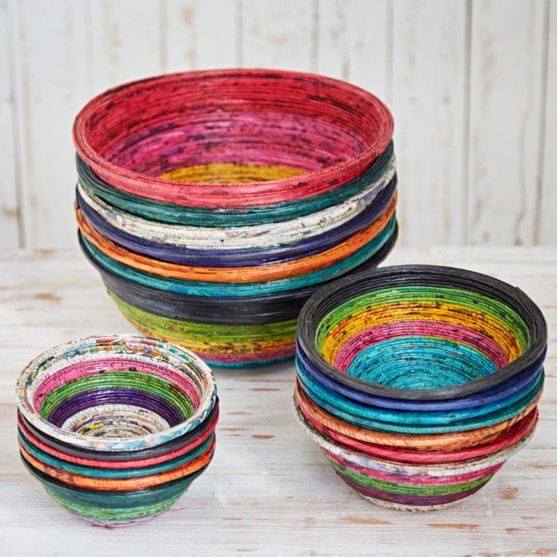 Three stacks of Round Recycled Newspaper Bowls in various sizes and vibrant colors are arranged on a light wooden surface, set against a white wooden wall.
