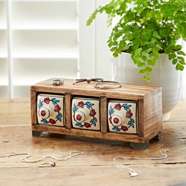 A White Floral Mango Wood 3 Drawer Jewellery Box sits on a wooden table beside a green potted plant. Jewelry items are spread out around the box.