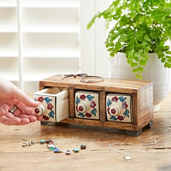A hand opens one of the three decorated small drawers in a White Floral Mango Wood 3 Drawer Jewellery Box. Jewelry pieces and a green plant are on the wooden surface nearby.