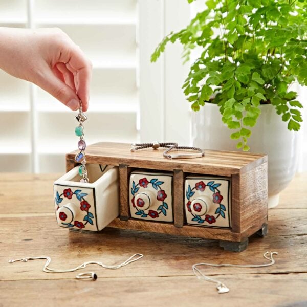 A hand places jewelry into one of the floral-patterned drawers of a White Floral Mango Wood 3 Drawer Jewellery Box. The mango wood jewellery box sits on a table next to a plant in a white pot, with jewelry items scattered on the table.