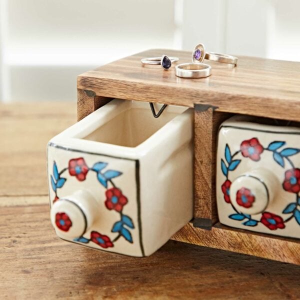 A White Floral Mango Wood 3 Drawer Jewellery Box with small ceramic drawers featuring white floral designs. One drawer is open, and three rings with different gemstones are displayed on top of the box.