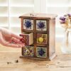 A hand opens a small drawer in the Multicoloured Mango Wood 6 Drawer Jewellery Box. Jewelry items are placed around the box on a wooden surface, and there is a vase with dried flowers in the background.