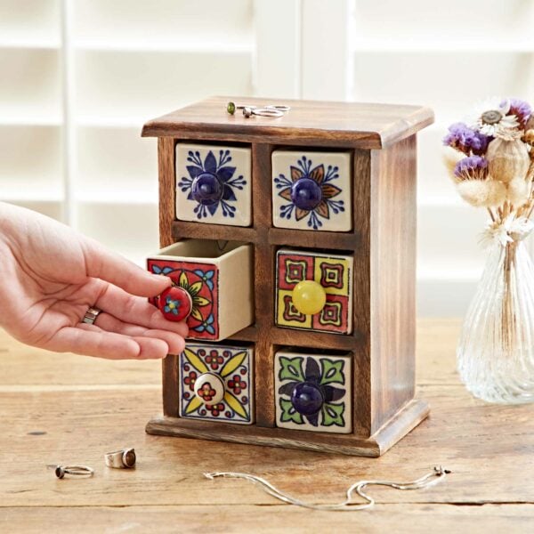 A hand opens a small drawer in the Multicoloured Mango Wood 6 Drawer Jewellery Box. Jewelry items are placed around the box on a wooden surface, and there is a vase with dried flowers in the background.