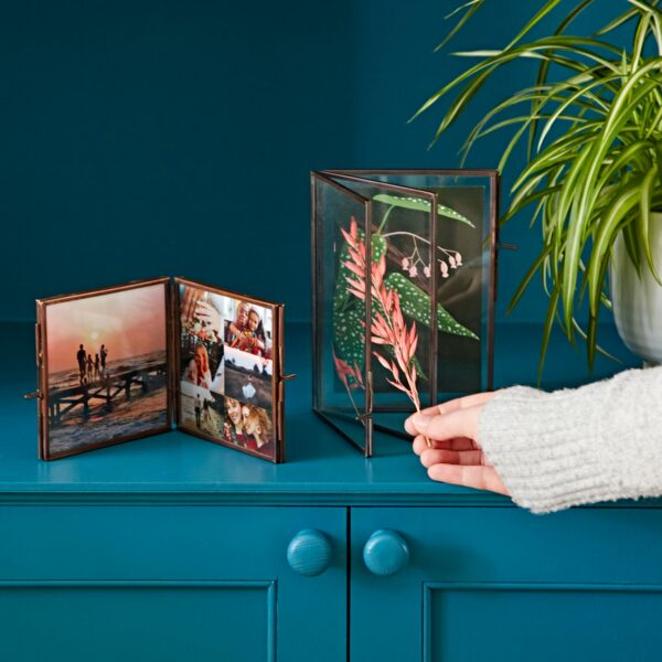 A hand in a gray sleeve adjusts an Antique Brass Recycled Glass Folded Double Photo Frame that showcases various photos and a leaf print. The double photo frame is placed on a teal cabinet next to a potted plant, giving the space an elegant touch.