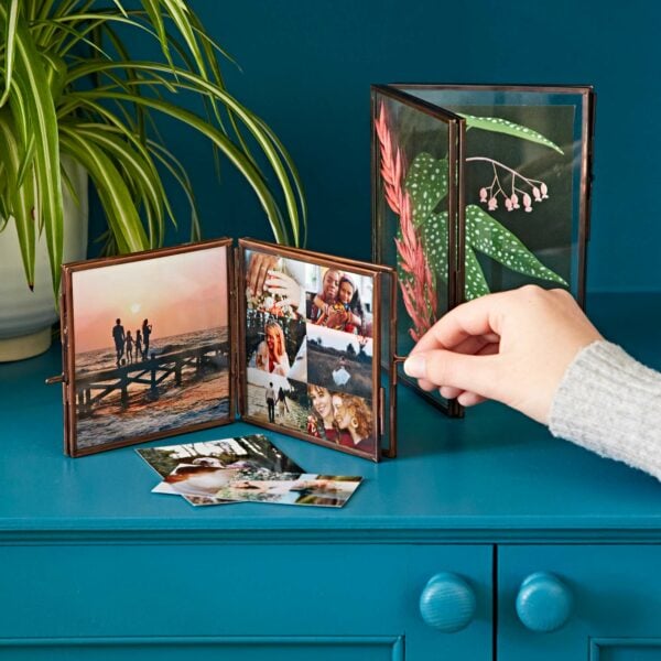 A hand adjusts an Antique Brass Recycled Glass Folded Double Photo Frame on a blue table, displaying various family photos and botanical prints. A plant in a white pot is in the background.