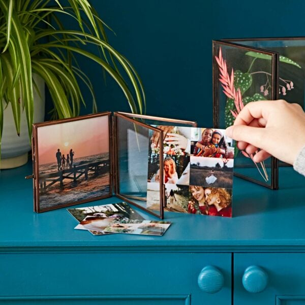 A hand adds a photo to an Antique Brass Recycled Glass Folded Double Photo Frame on a blue cabinet. Several photos, including a sunset and people smiling, are visible. A green plant is in the background.