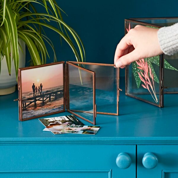 A hand adjusts an Antique Brass Recycled Glass Folded Double Photo Frame on a blue dresser. One section displays a family photo at a pier, with loose photos lying nearby. A green plant is in the background.