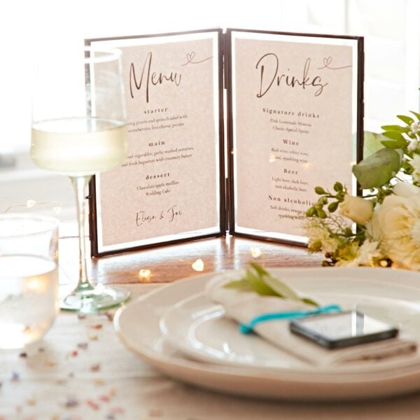 A wedding reception setup with a menu and drinks list, featuring a wine glass, a decorated plate with a smartphone, floral arrangement, string lights, and an Antique Brass Recycled Glass Folded Double Photo Frame.