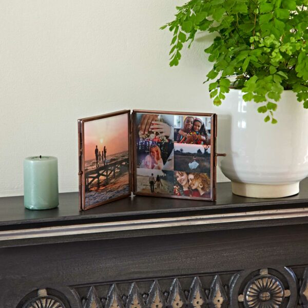 A small Antique Brass Recycled Glass Folded Double Photo Frame displaying multiple pictures sits on a mantelpiece next to a green candle and a potted plant.