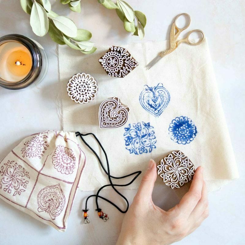 A hand holds a carved stamp above blue-stamped fabric. Nearby are Four Assorted Design Wooden Printing Blocks, small scissors, a patterned pouch, a candle, and greenery.