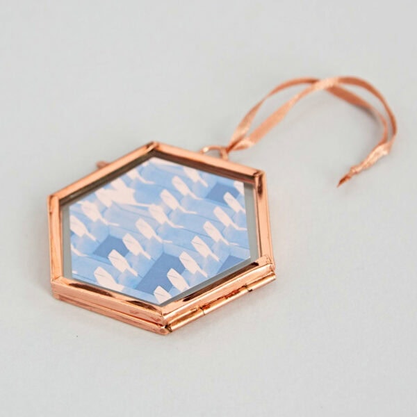 A hexagonal ornament with a rose gold frame and ribbon, featuring a geometric pattern of white shapes on a blue background. This elegant Alia Mini Glass Hexagon doubles as a hanging photo frame, perfect for showcasing cherished memories.