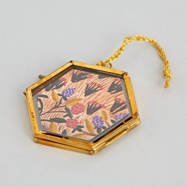 An ornate hexagonal gold locket with a floral pattern on the front, featuring a braided golden chain, rests on a plain surface next to an Alia Mini Glass Hexagon Hanging Photo Frame.
