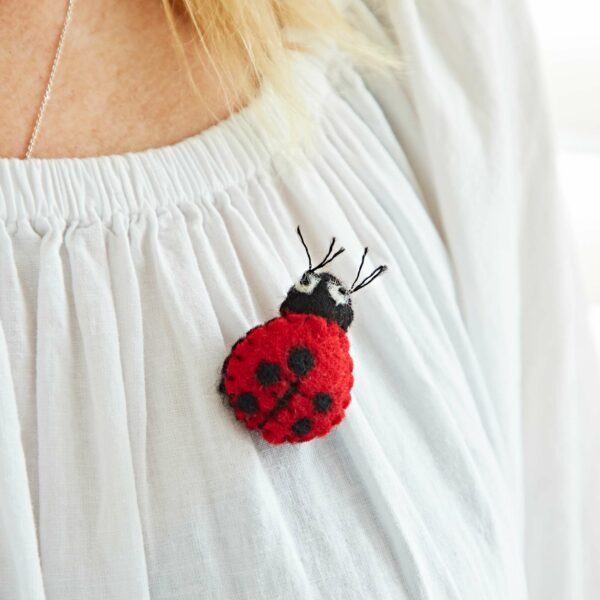 A close-up of a Pack of 3 Felt Ladybird Brooches with black dots, pinned on a woman's white blouse with gathered neckline detail.