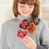 A person with long hair and bangs wears a grey turtleneck sweater and holds a pink and yellow handmade yarn flower, alongside three other similar flowers pinned to their sweater. They also showcase a Pack of 3 Felt Butterfly Brooches, adding an extra touch of charm to the ensemble.