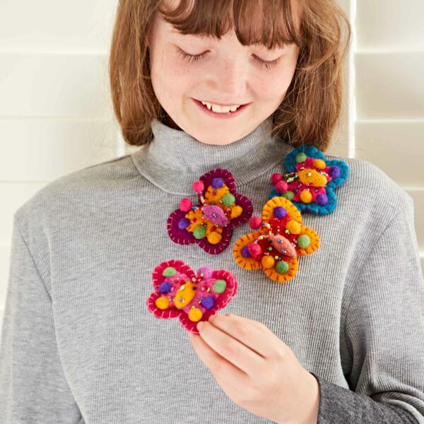 A person with long hair and bangs wears a grey turtleneck sweater and holds a pink and yellow handmade yarn flower, alongside three other similar flowers pinned to their sweater. They also showcase a Pack of 3 Felt Butterfly Brooches, adding an extra touch of charm to the ensemble.