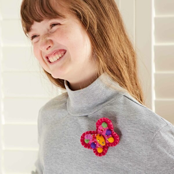 Young person with long brown hair wears a gray turtleneck and smiles broadly, displaying one of their Pack of 3 Felt Butterfly Brooches on their shirt. This stunning piece is part of a delightful pack of 3, each handcrafted and bursting with vibrant colors.