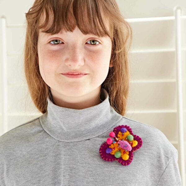 A girl with long brown hair is wearing a grey turtleneck sweater and a colorful embroidered brooch shaped like a flower from the Pack of 3 Felt Butterfly Brooches.