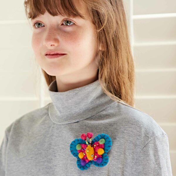 A person wearing a grey turtleneck sweater with a colorful, Pack of 3 Felt Butterfly Brooches pinned to it.