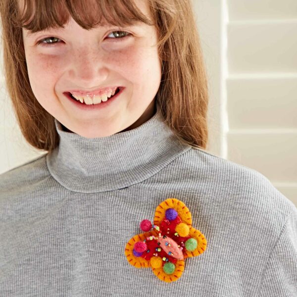 A person with long hair smiles, wearing a gray turtleneck sweater and a colorful felt brooch in the shape of a butterfly adorned with pom-poms and beads. Consider accessorizing with the Pack of 3 Felt Butterfly Brooches to add more vibrant touches to your outfits.