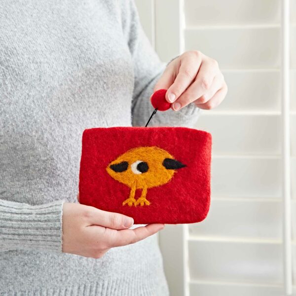 Person holding a small Felt Bird Purse with a cartoon bird design. The pouch, resembling a mini purse, has a round red zipper pull, and the person is wearing a gray sweater. Background includes white window shutters.