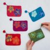 Five colorful Felt Flower Purses with detailed floral patterns sit on a white surface. Someone is holding the green purse, featuring a playful pom-pom zipper.