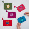 A set of five vibrant Felt Daisy Purses with zip closures is showcased on a light surface, with the blue purse held in hand.