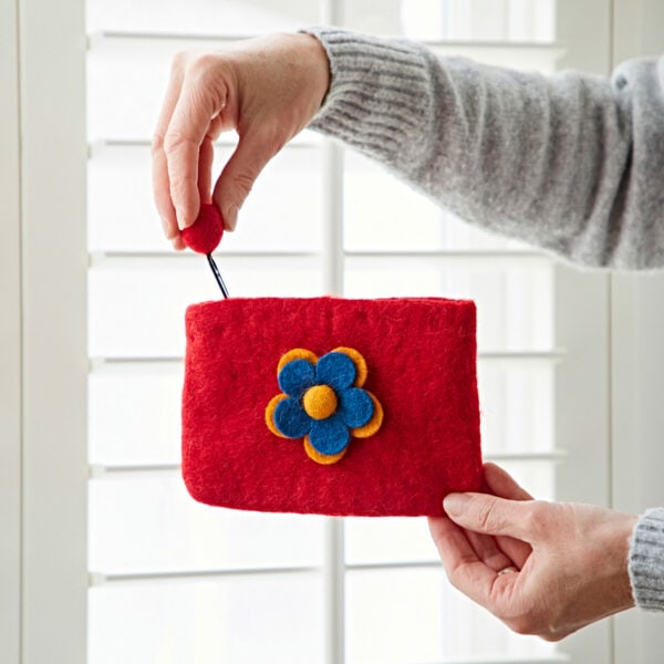 A pair of hands unzips the vibrant Felt Daisy Purse with a round pull, set against a backdrop of white shutters.