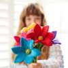 A person holding a bouquet of colorful felt flowers featuring the Felt Lily Flower in front of a blurred background with window shutters.
