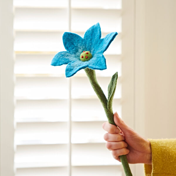 Someone holds a large Felt Lily Flower, featuring a blue bloom with a green stem and leaves, in front of a window with white blinds.