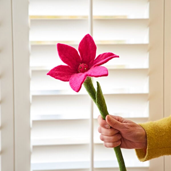 A hand holds a Felt Lily Flower with a pink blossom and green stem in front of white window shutters.