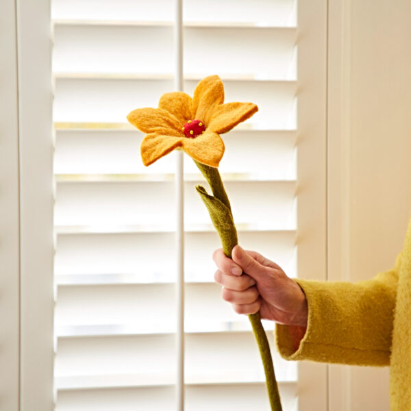 A person holds the Felt Lily Flower, featuring a long green stem and bright yellow petals, in front of white window shutters.
