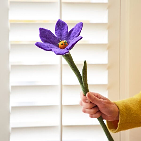 A hand holds a large Felt Lily Flower, featuring a purple hue and a yellow center, in front of white window shutters.