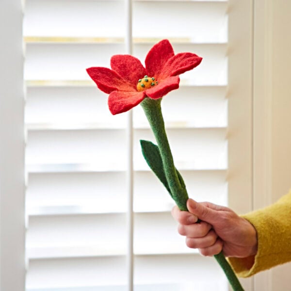 A hand presents a Felt Lily Flower, featuring a red bloom and a long green stem, before white window shutters.