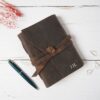 A Personalised Buffalo Leather Wraparound Journal featuring embossed initials "J.H." is displayed on a white wooden surface, alongside a blue pen and red dried flowers.
