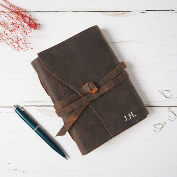 A Personalised Buffalo Leather Wraparound Journal featuring embossed initials "J.H." is displayed on a white wooden surface, alongside a blue pen and red dried flowers.