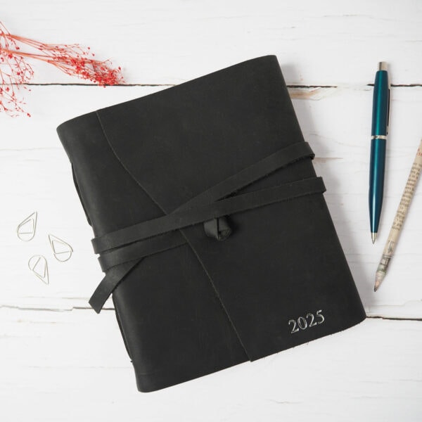 The Personalised Large Buffalo Leather Wraparound Journal, featuring "2025" embossed on its cover, is secured with a thin strap and laid out on a white wooden surface. Accompanying it are a blue pen, pencil, and paper clips.