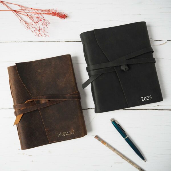 Two Personalised Large Wraparound Journals are elegantly displayed on a white wooden surface. One journal, crafted from buffalo leather, has "14.02.25" embossed on its cover, while the other is a sleek black design featuring the year "2025." Nearby, dried flowers accentuate the scene along with a pen and pencil.