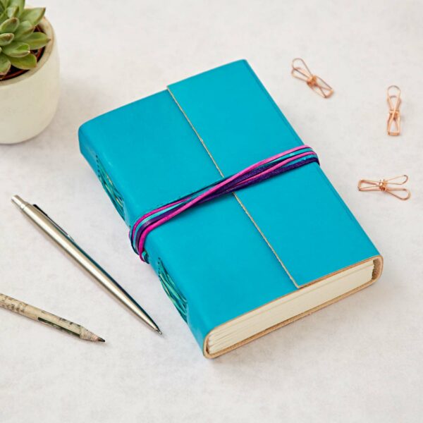 A closed Personalised Coloured Distressed Leather 3-String Journal with vibrant strings, accompanied by a pen, pencil, rose gold clips, and a small potted succulent on a light surface.