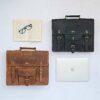A Personalised Buffalo Leather Vintage Satchel sits beside an open book with glasses, a closed silver laptop, and another leather briefcase on a light surface.