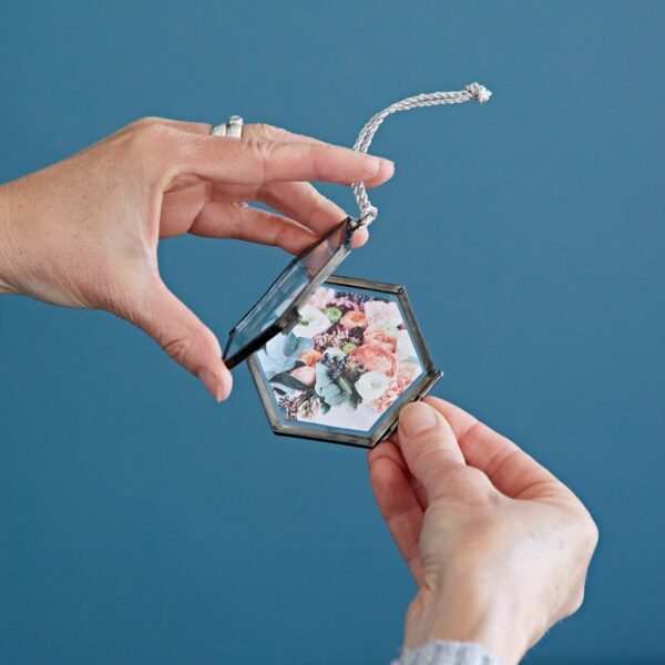 Person holding an Alia Mini Glass Hexagon Hanging Photo Frame with a picture of flowers inside against a blue background.