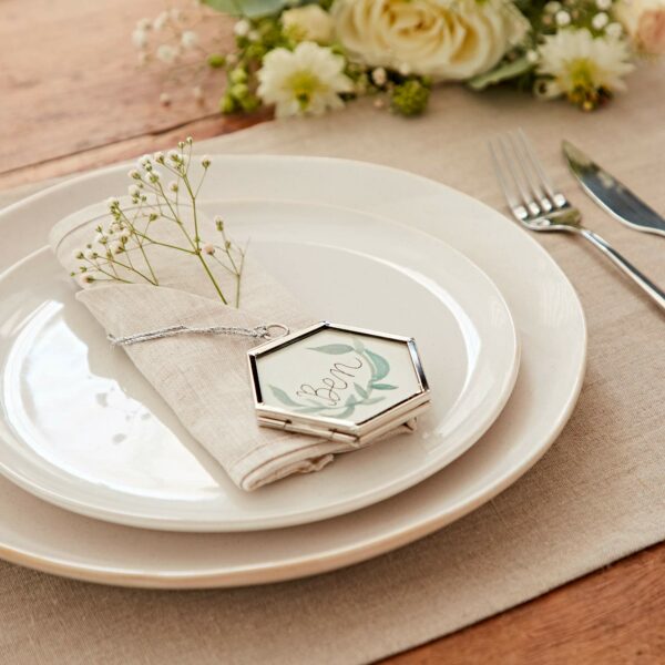 A table setting with a white plate, beige napkin, silverware, and a hexagonal locket inscribed with "love," complemented by an Alia Mini Glass Hexagon Hanging Photo Frame. Floral arrangement in the background.