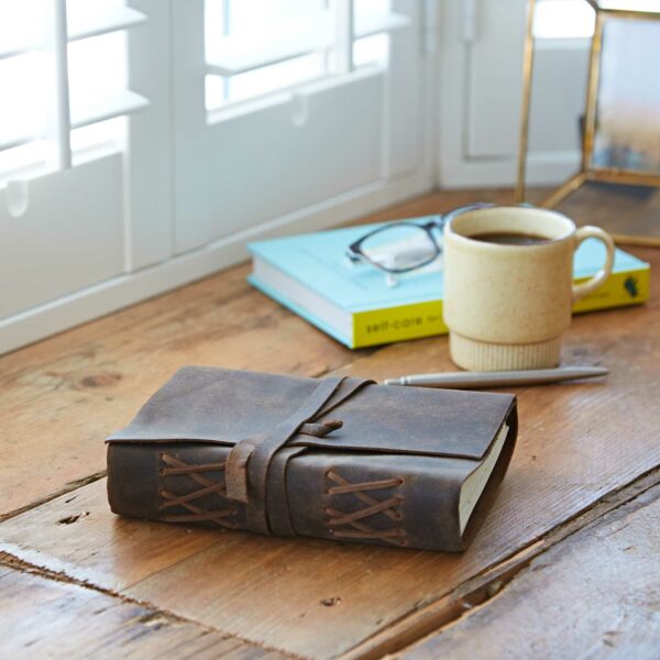 A Personalised Buffalo Leather Wraparound Journal, a pen, a beige mug of coffee, and a book with glasses on top are arranged on a wooden table by a window with shutters.