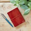 A naturally dyed, handmade Personalised Mini Leather Pocket Notebook with a pen and a plant on a wooden table.