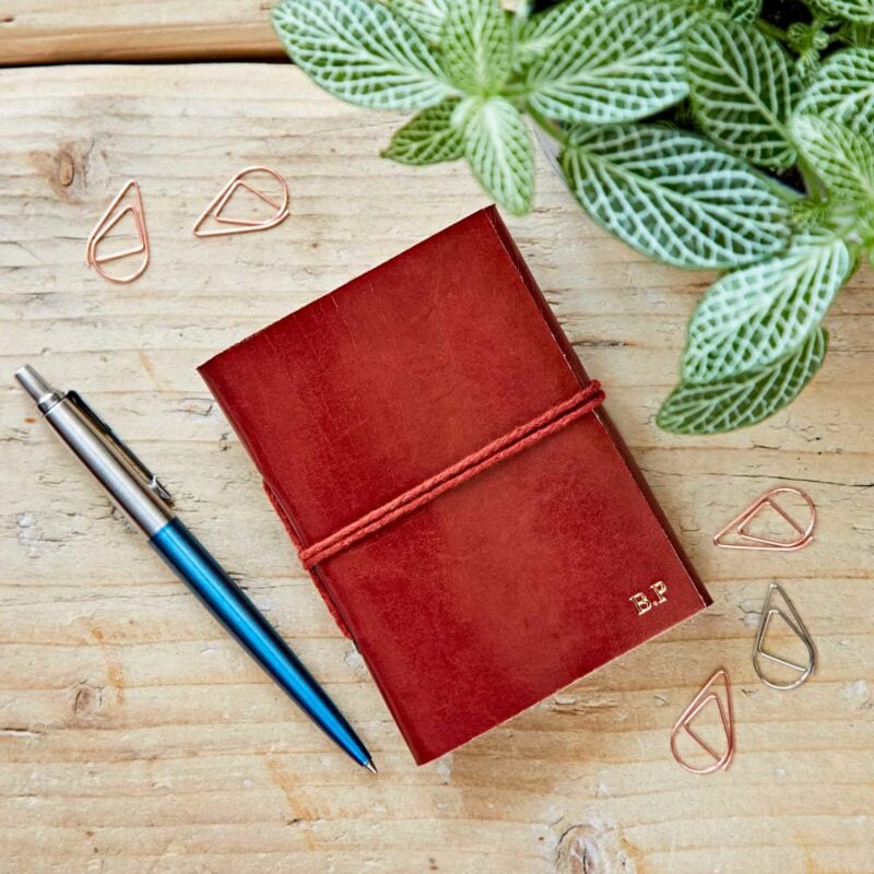 A naturally dyed, handmade Personalised Mini Leather Pocket Notebook with a pen and a plant on a wooden table.