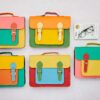 Five Multicoloured Recycled Leather Satchels with buckle fastenings are displayed on a light surface beside a book and spectacles.