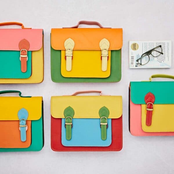 Five Multicoloured Recycled Leather Satchels with buckle fastenings are displayed on a light surface beside a book and spectacles.