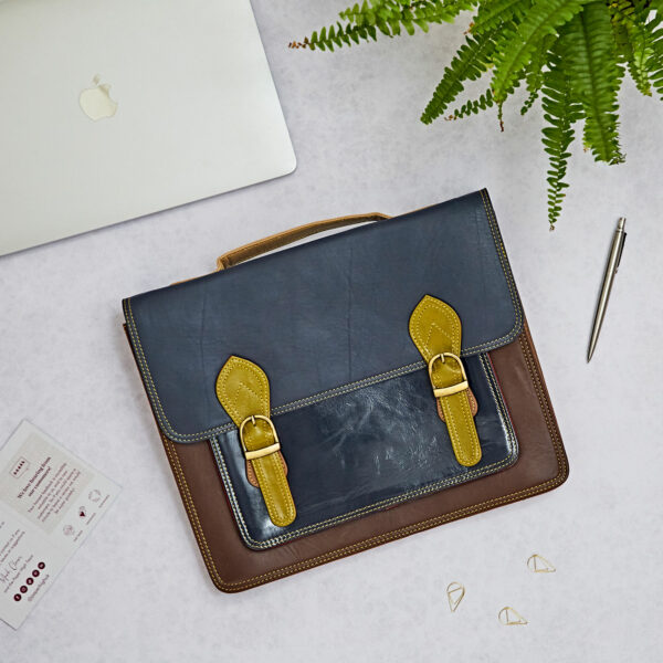 The Multicoloured Recycled Leather Satchel rests on a white surface alongside a laptop, a fern, a pen, paperclips, and paperwork.