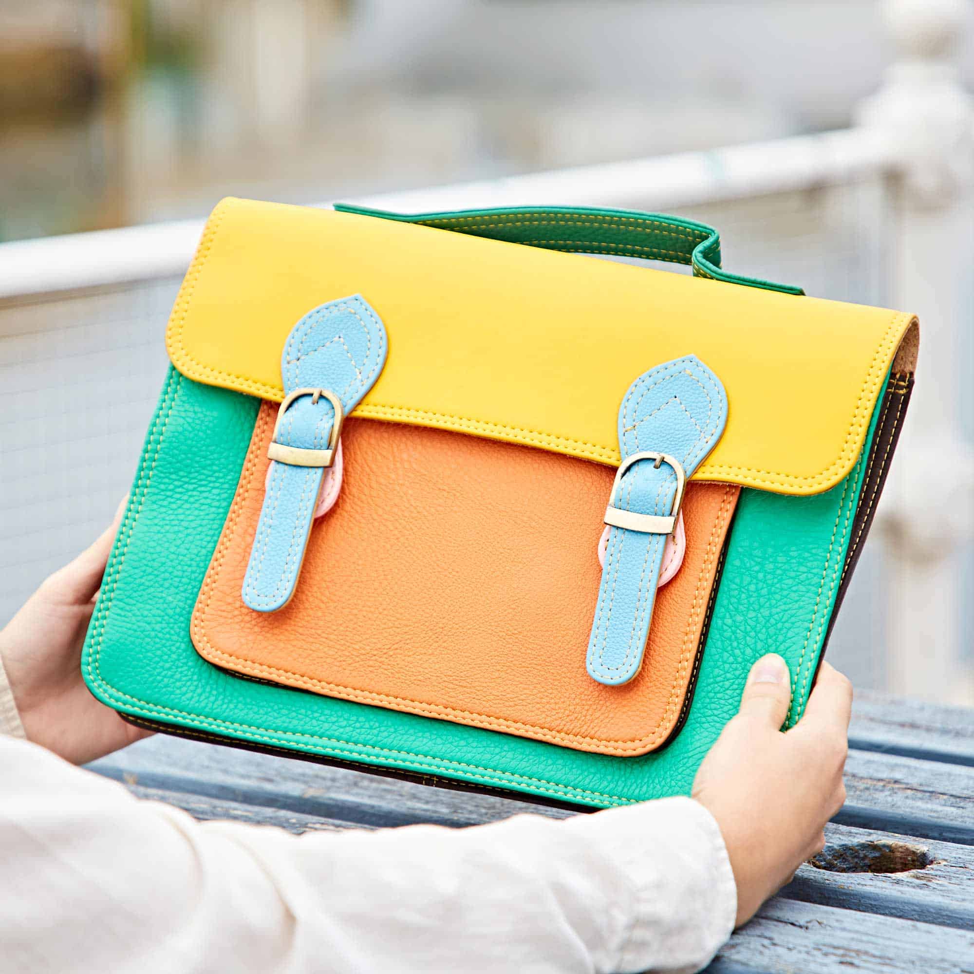 A person holds the Multicoloured Recycled Leather Satchel, featuring a yellow flap, blue straps, an orange front pocket, and green sides.