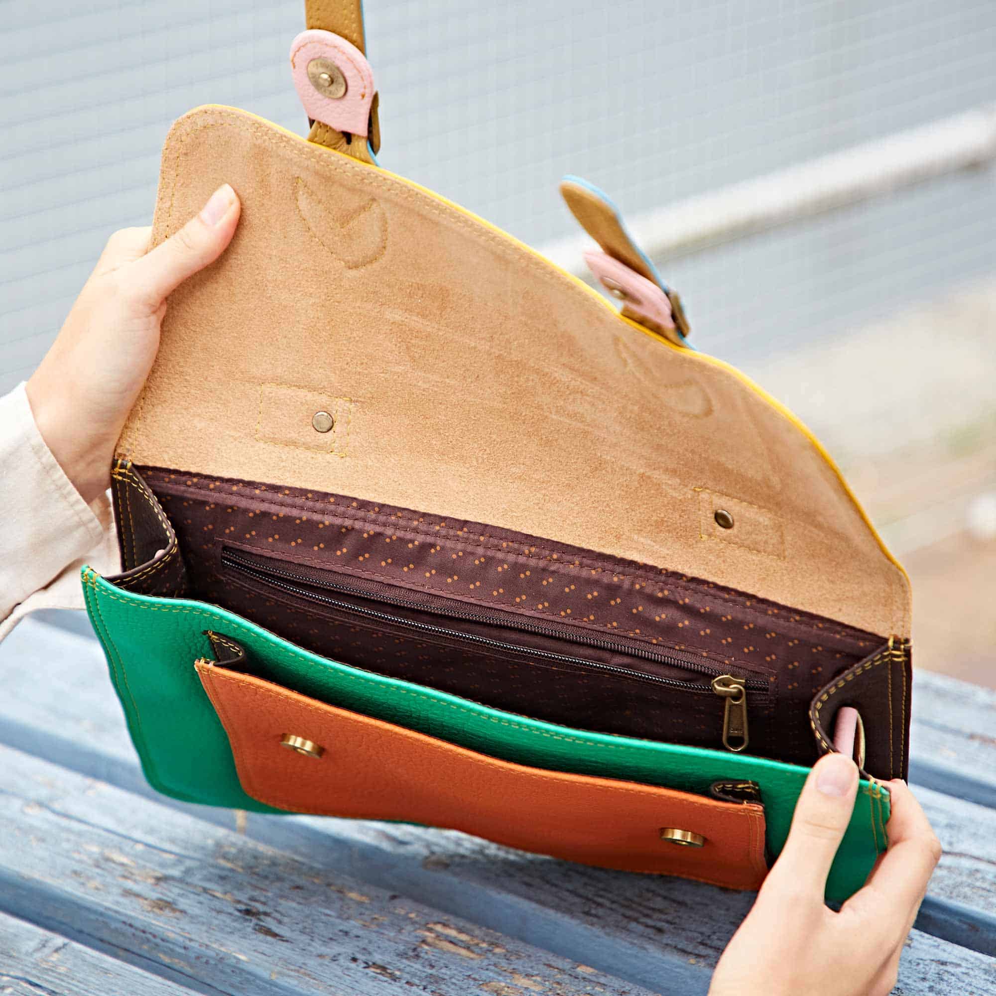 A person opens the Multicoloured Recycled Leather Satchel to reveal its vibrant suede interior, zip-fastened compartment, and multiple pockets.