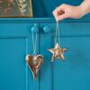 A hand holds **Mango Wood Heart and Star Decorations**, both hanging from strings on a blue cabinet.